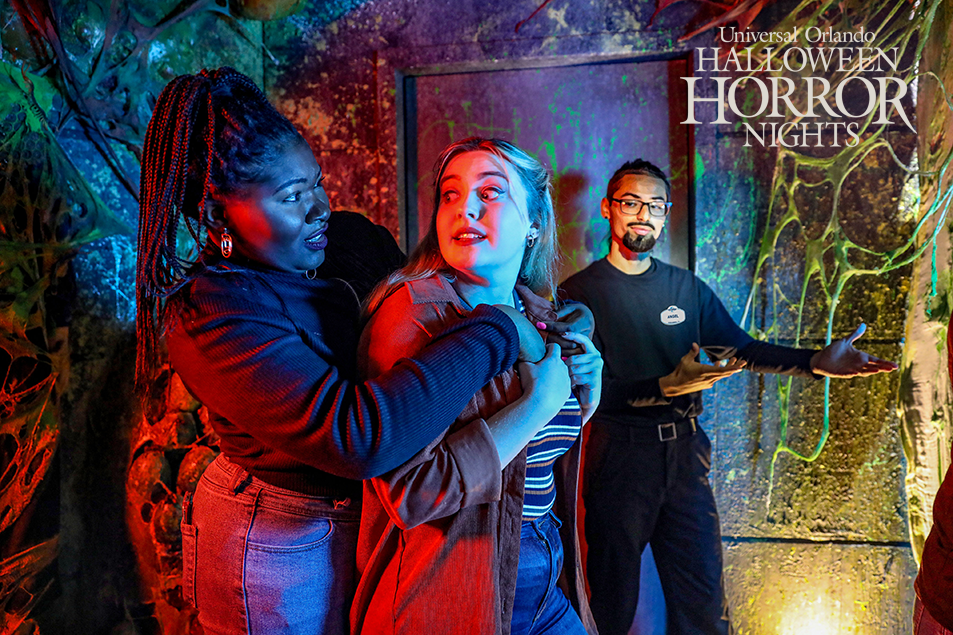Two woman are holding on to each other while walking through a Haunted House, they are scared. There is a man in the background who is guiding them. In the top right logo there is a logo that says Universal Studios Halloween Horror Nights.