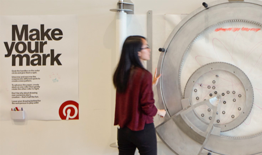 Woman standing in front of an extra large spirograph and a poster on the wall reads, Make Your Mark.