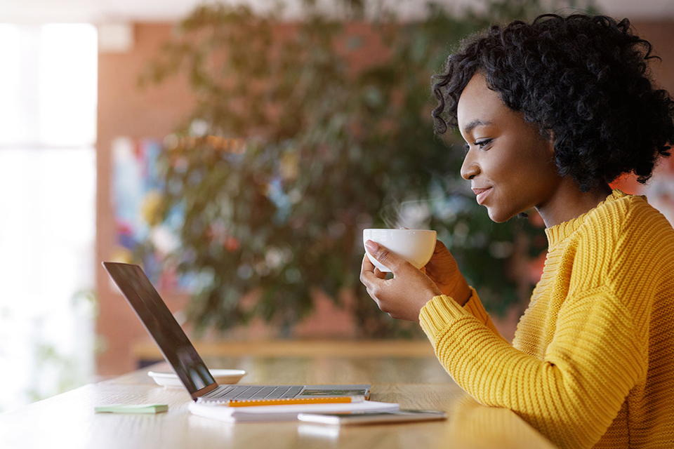 Woman sipping from a coffee cup while smiling and looking at her laptop
