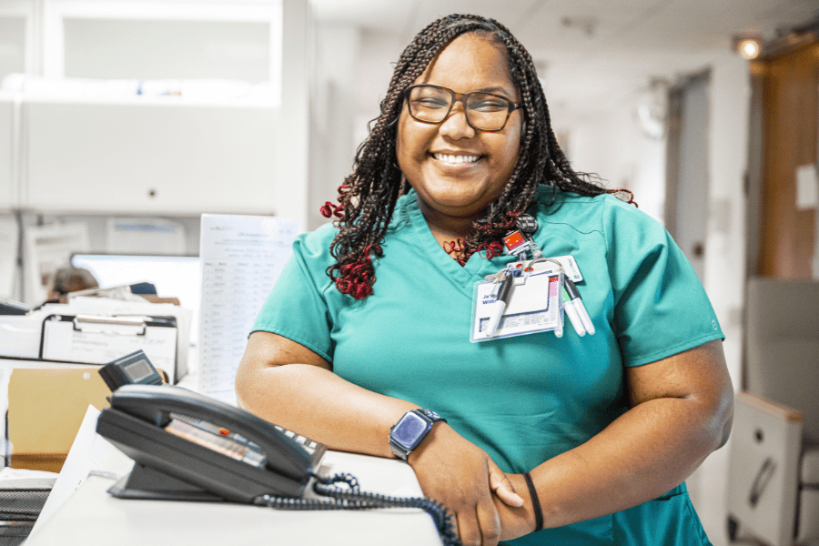 Smiling Nurse Support staff member stands by a phone at a nurse station.