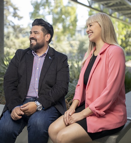 Two professionals smiling and sitting down in a bench with trees and bushes in the background.