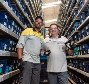 Two male CNH Supply Chain employees stand together in a product warehouse and smile at the camera.