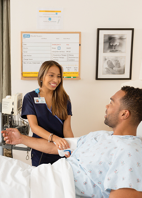 A smiling patient in a hospital bed with a nurse in blue scrubs caring for him, surrounded by medical equipment.