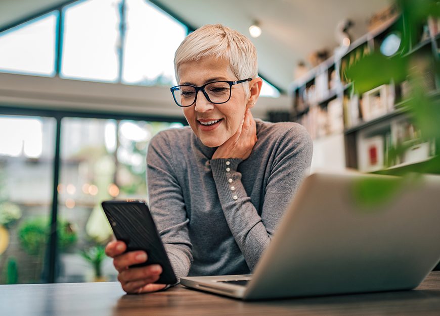 Mature woman in her home with a laptop in front of her and looking at her phone.