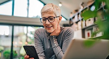 Mature woman in her home with a laptop in front of her and looking at her phone.