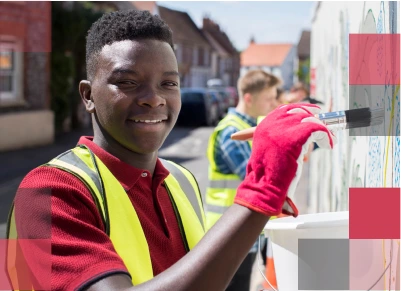 A smiling male African American high school student and other students working on painting a wall outside