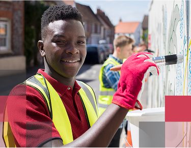 A smiling male African American high school student and other students working on painting a wall outside