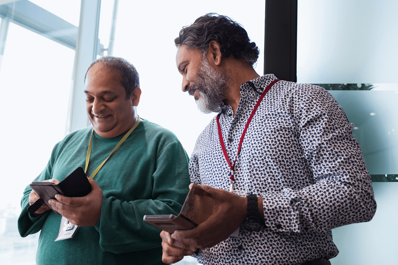 Two individuals conversing while looking at their mobile devices in an office. One is wearing a green sweater and the other is wearing a patterned shirt and has a beard and mustache.