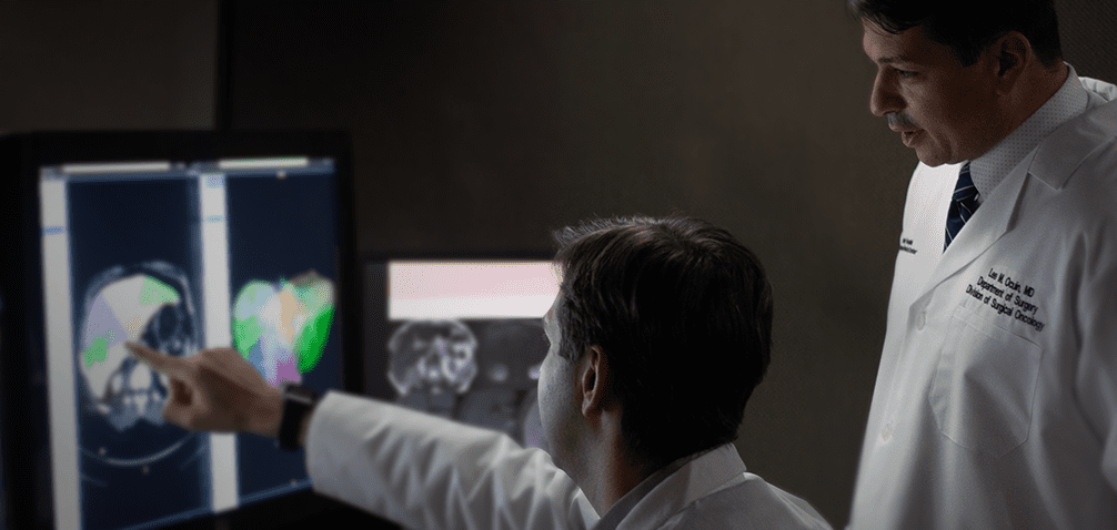 Two doctors in white coats analyze and discuss medical imaging scans on a computer screen in a dimly lit room.