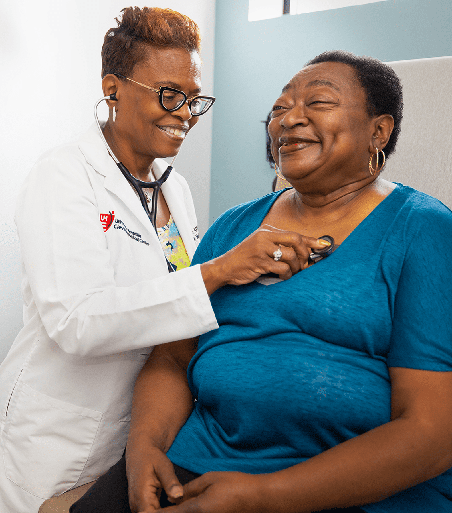 A smiling healthcare professional in a white coat uses a stethoscope to check a happy patient in a clinical setting.
