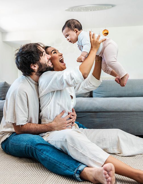 A smiling mother and father sitting on the floor with the mother holding their child in the air