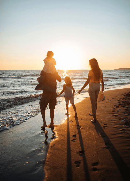 A family of four walks along the beach at sunset, with one child on an adult’s shoulders and the other holding hands while waves reach the shore.