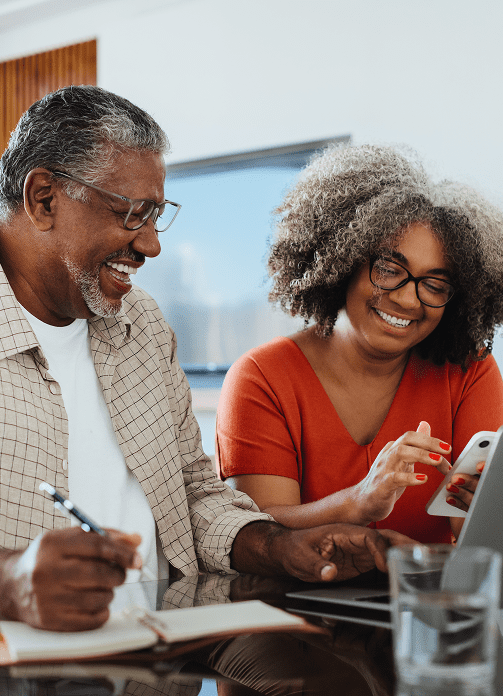 An older man and woman smile while sitting together at a table, looking at a smartphone and laptop with a notebook and pen nearby.