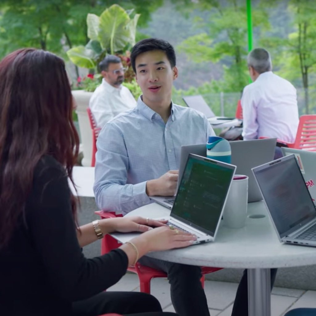 Two people seated at a round outdoor table with laptops, engaged in conversation. They are surrounded by others at similar tables in a patio area with greenery and trees in the background.