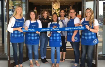 A diverse team of Bath and Body Works employees wearing their uniformed blue aprons holding a blue ribbon together in front of a new store location. 