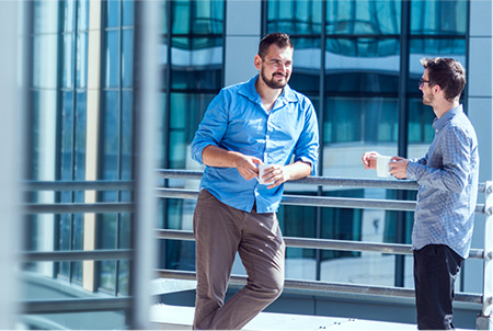 Two men engaged in a casual conversation, as they stand outside of their local ADP office building on a sunny day.