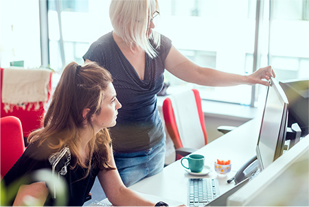 Two women work together in a modern office space. One sits and the other stands as they look at information on a desktop computer monitor.
