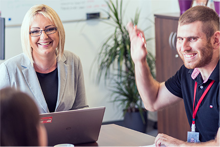 Employees sit together at a square table. The smiling woman at the head of the table has her laptop open, as the man next to her smiles and lifts his arm in a high five type of gesture.