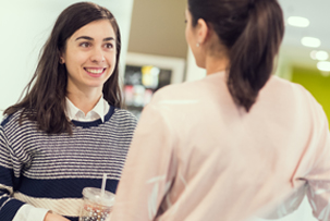 Two women having a casual talk while standing in an office environment. The woman on the left is holding a beverage in a clear cup with a straw.