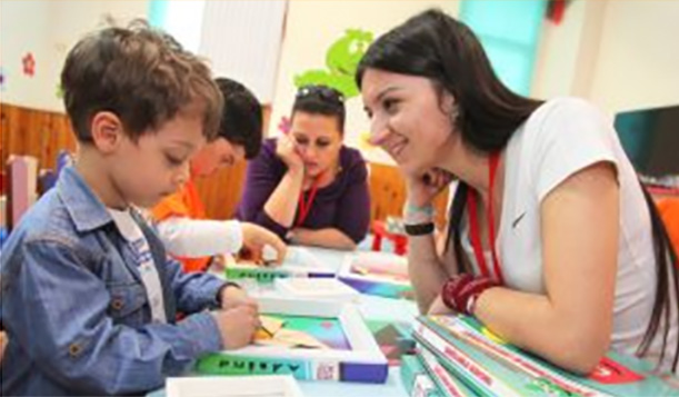 Dwie wolontariuszki siedzą przy stole, pomagając dwóm chłopcom w czytaniu i innych zajęciach edukacyjnych.