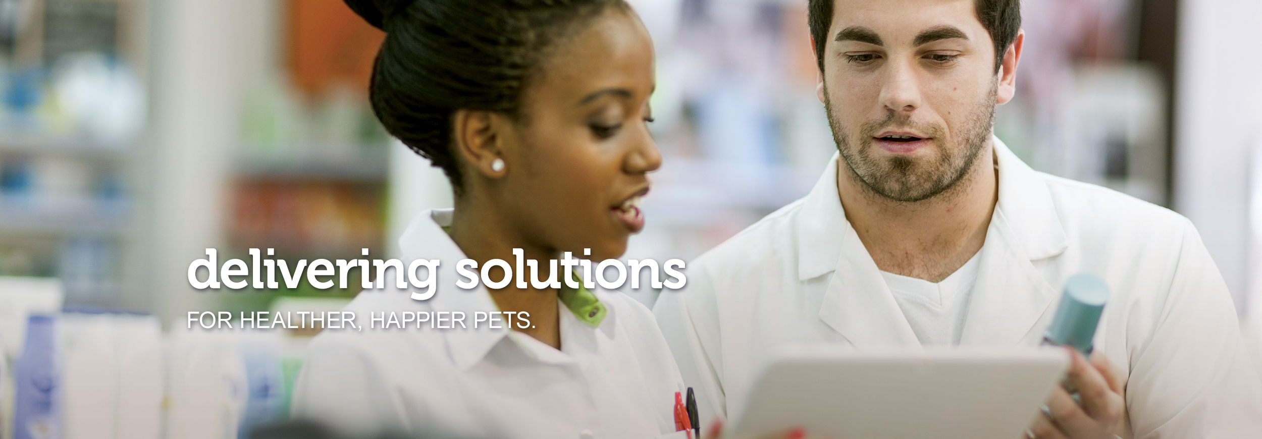 A male and a female Pharmacist confer over a clipboard and medications to find the correct prescription. Text across the image reads, “delivering solutions for healthier, happier pets.”