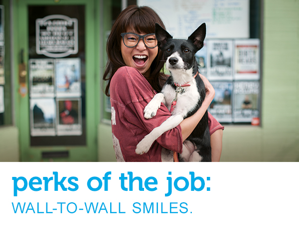 Excited young woman in glasses holds her equally happy-looking black and white dog as they look at the camera. Text across the image reads, “perks of the job: wall-to-wall smiles.”