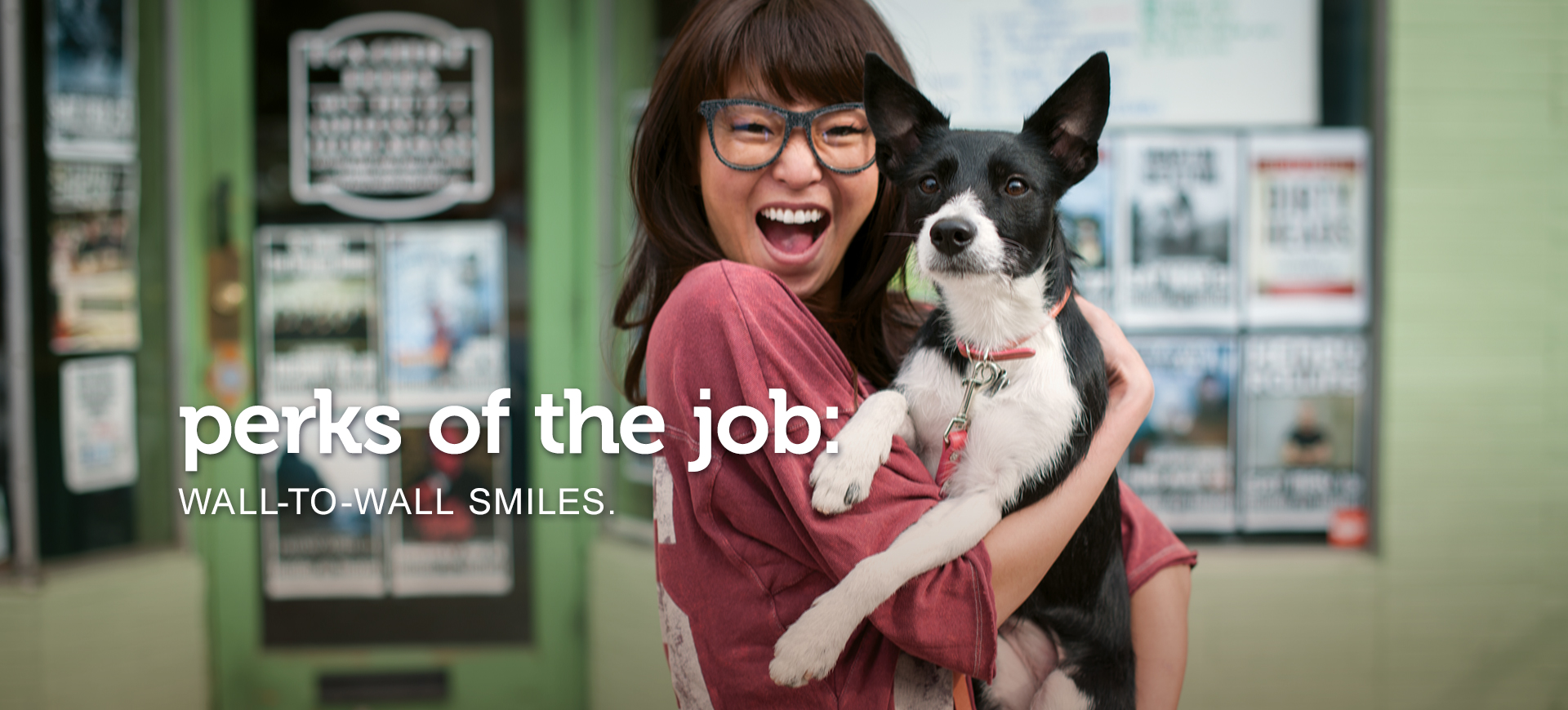 Excited young woman in glasses holds her equally happy-looking black and white dog as they look at the camera. Text across the image reads, “perks of the job: wall-to-wall smiles.”