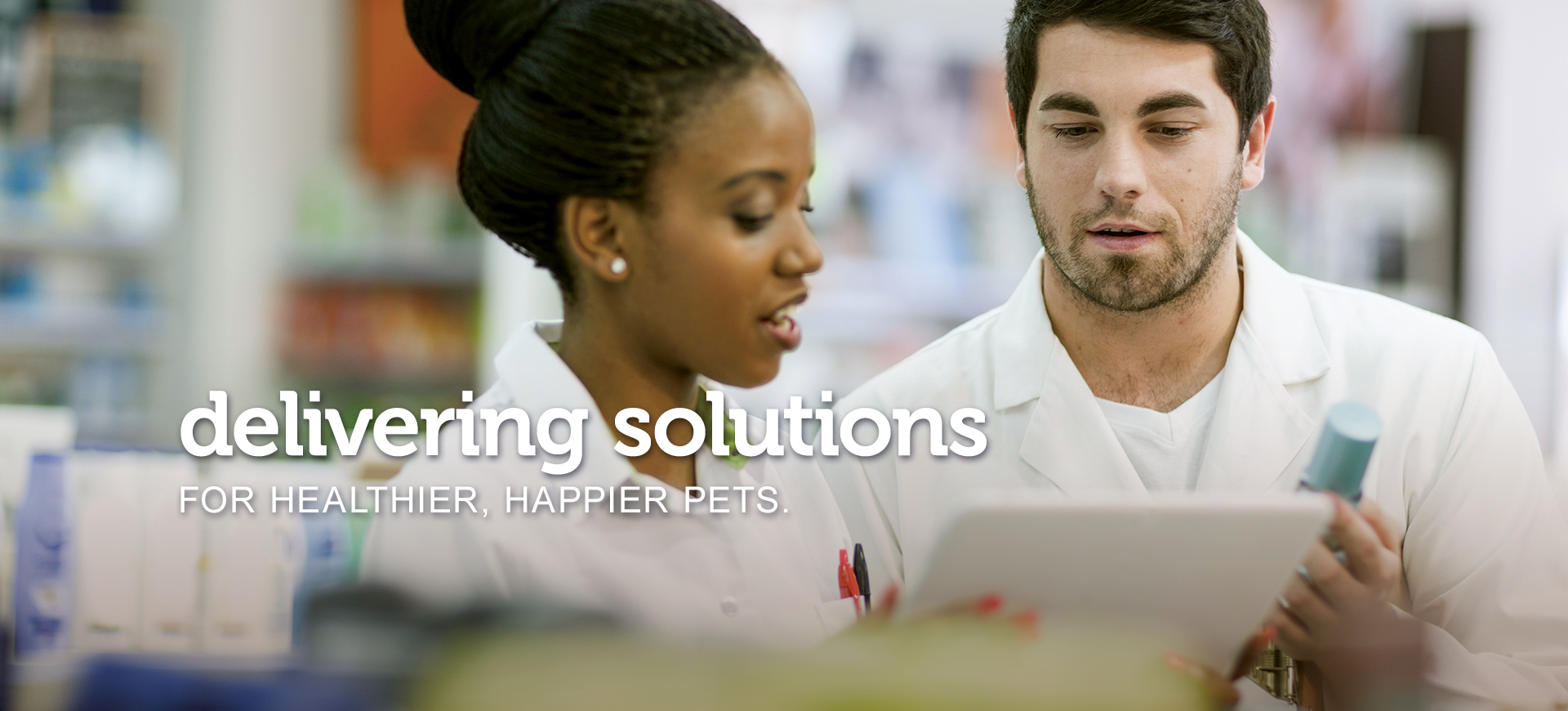 A male and a female Pharmacist confer over a clipboard and medications to find the correct prescription. Text across the image reads, “delivering solutions for healthier, happier pets.”