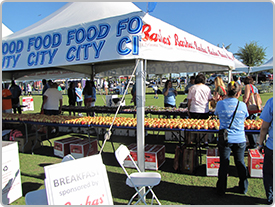 Línea de fruta fresca en una mesa debajo de una carpa blanca de Food City en un evento al aire libre de la comunidad local. La gente camina cerca de la carpa.