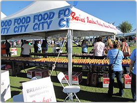 Línea de fruta fresca en una mesa debajo de una carpa blanca de Food City en un evento al aire libre de la comunidad local. La gente camina cerca de la carpa.