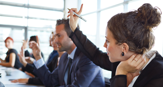 A group of adults in a conference room setting. One woman has her hand raised.