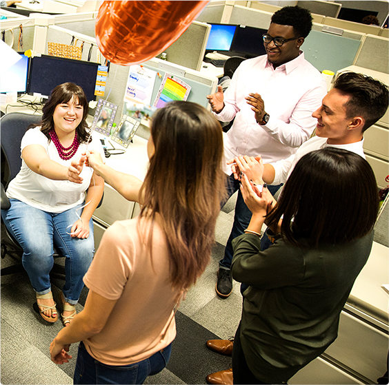 A group of male and female employees celebrating in an office cubicle, with a red balloon.